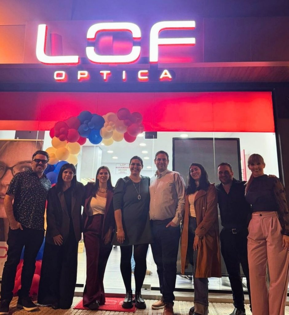 Group of eight adults posing outside a LOF Optica storefront with colorful balloon decorations for a grand opening.