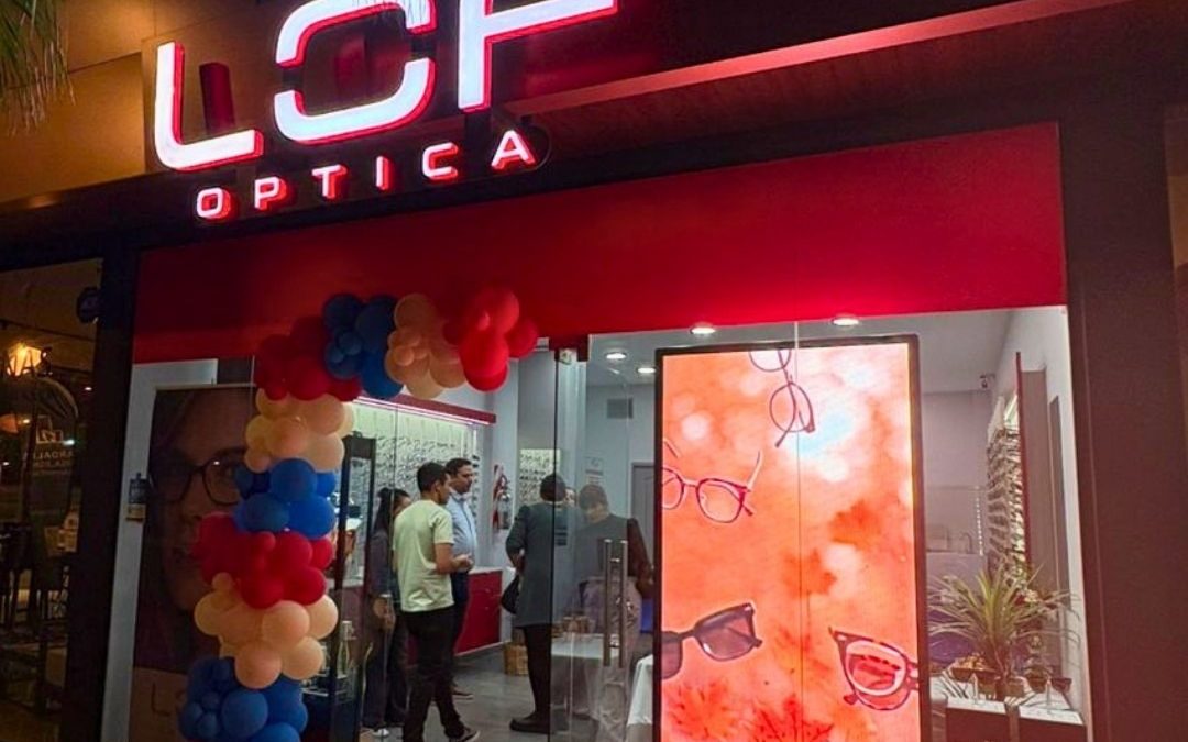 Storefront of LCF Optica at night with a red-lit sign, glass doors, and a colorful balloon arch; customers inside looking at eyewear.